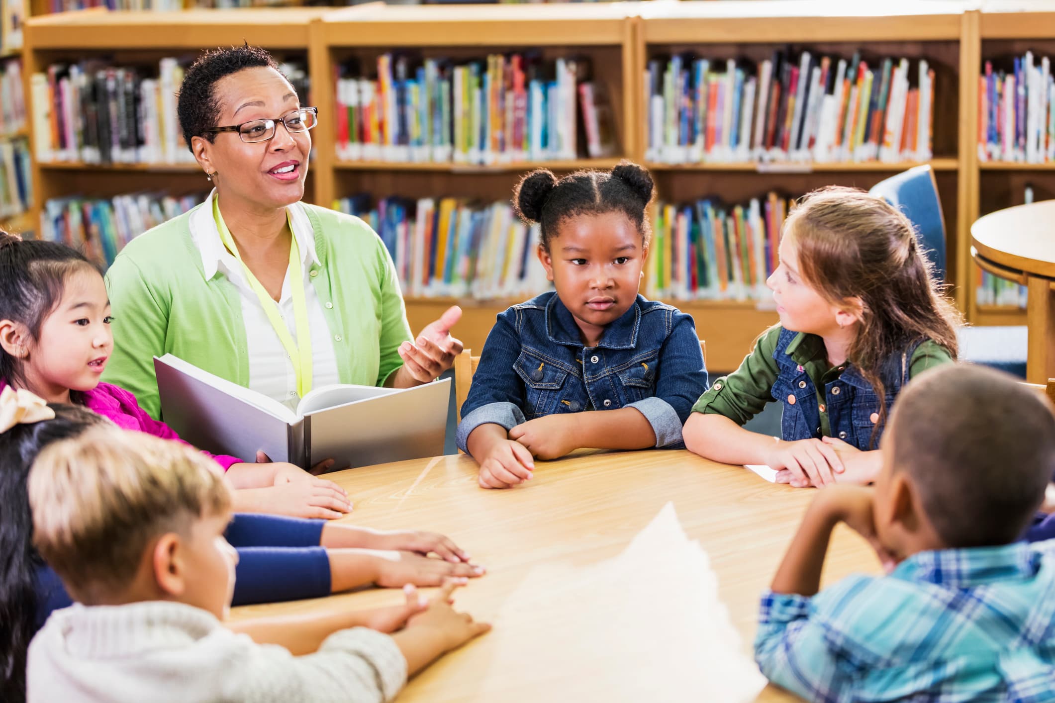 A multi-ethnic group of six children, 6 and 7 years old, sitting at a round table in the library with their teacher, a mature African-American woman, who is reading a book to them.