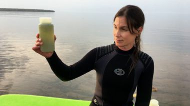 A woman in a wetsuit holding a clear plastic jar of algae and water